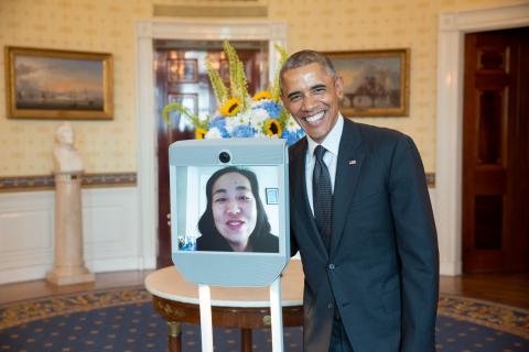 Photo of President Obama standing next to an electronic device with a viewscreen featuring the face of a smiling Asian American woman