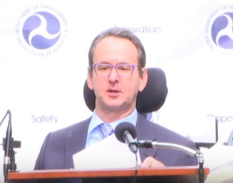 Image of Henry Claypool sitting in his wheelchair behind a podium and in front of a backdrop with US Department of Transportation logos